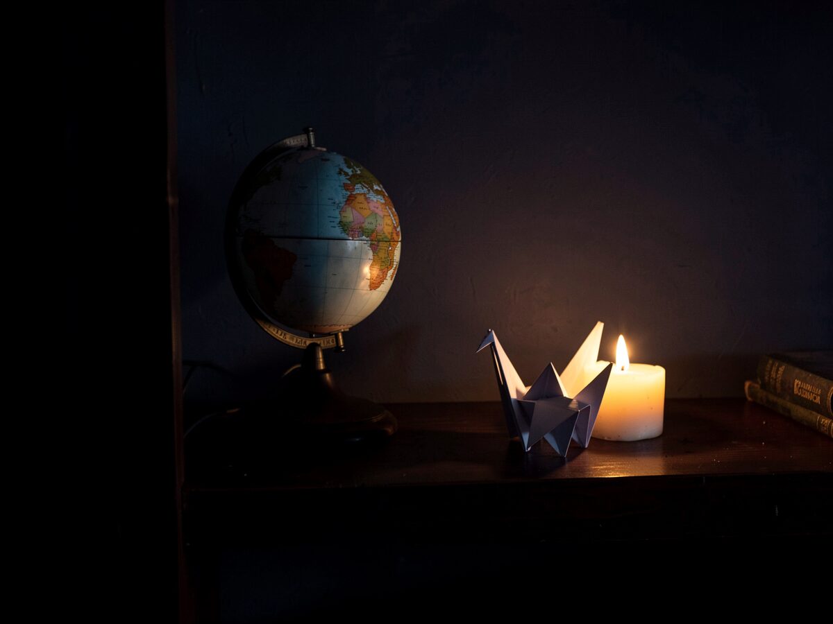 a desk globe and a folded paper crane beside a candle on a dark shelf, deep shadow, a faint warm glow, still life, no readable text