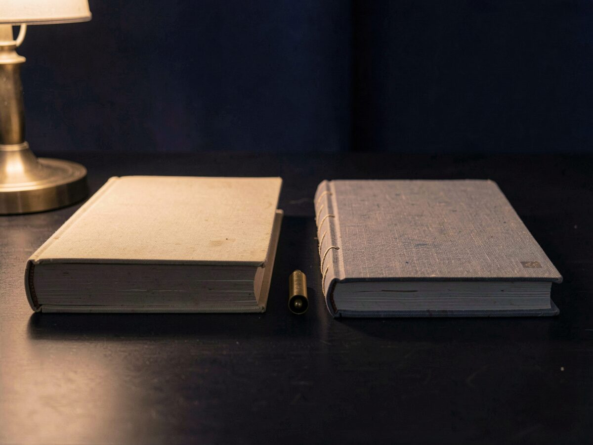 two clothbound manuals side by side on a dark desk, one with a plain western spine and one with a japanese-style stab binding, a small brass marker between them, no readable text