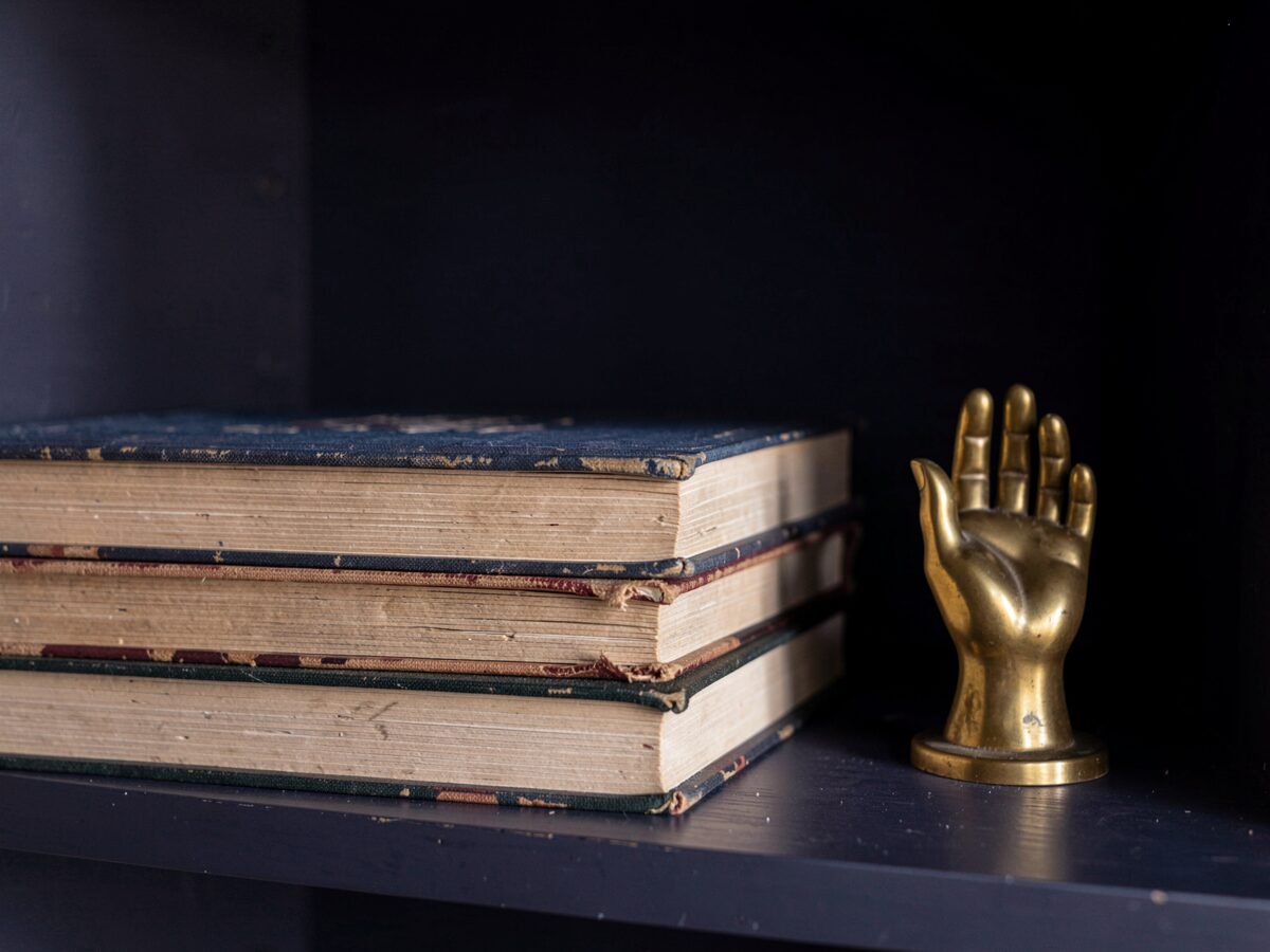 a short stack of old clothbound books seen from the page-edge side only, beside a small brass bookend shaped like an open hand, on a dark shelf in lamplight, dark indigo interior, photorealistic, no visible spines, no text