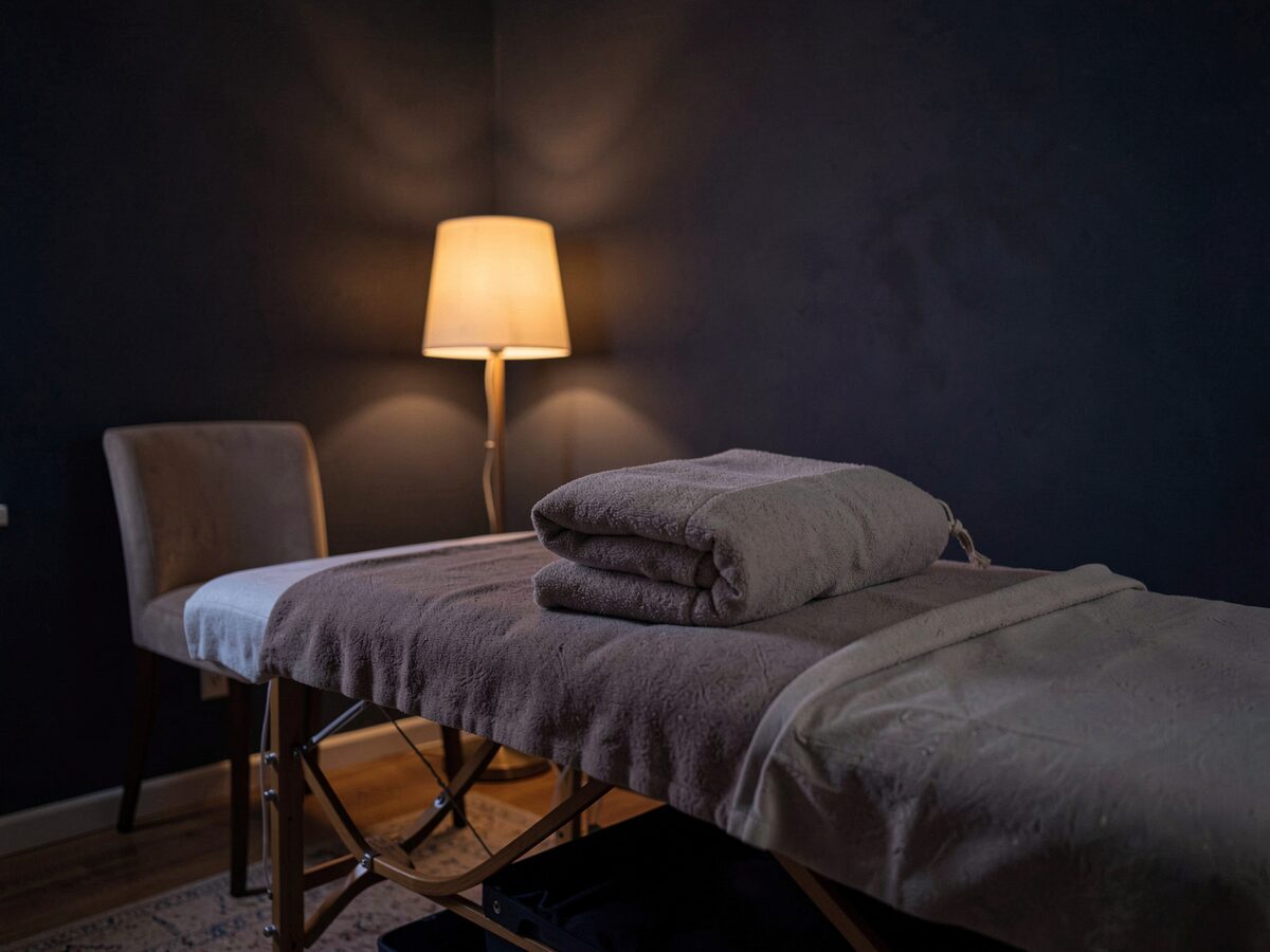 a massage table with a folded blanket and a small pillow in a dim quiet room, a chair beside it, warm lamp light against dark walls, no people, no readable text